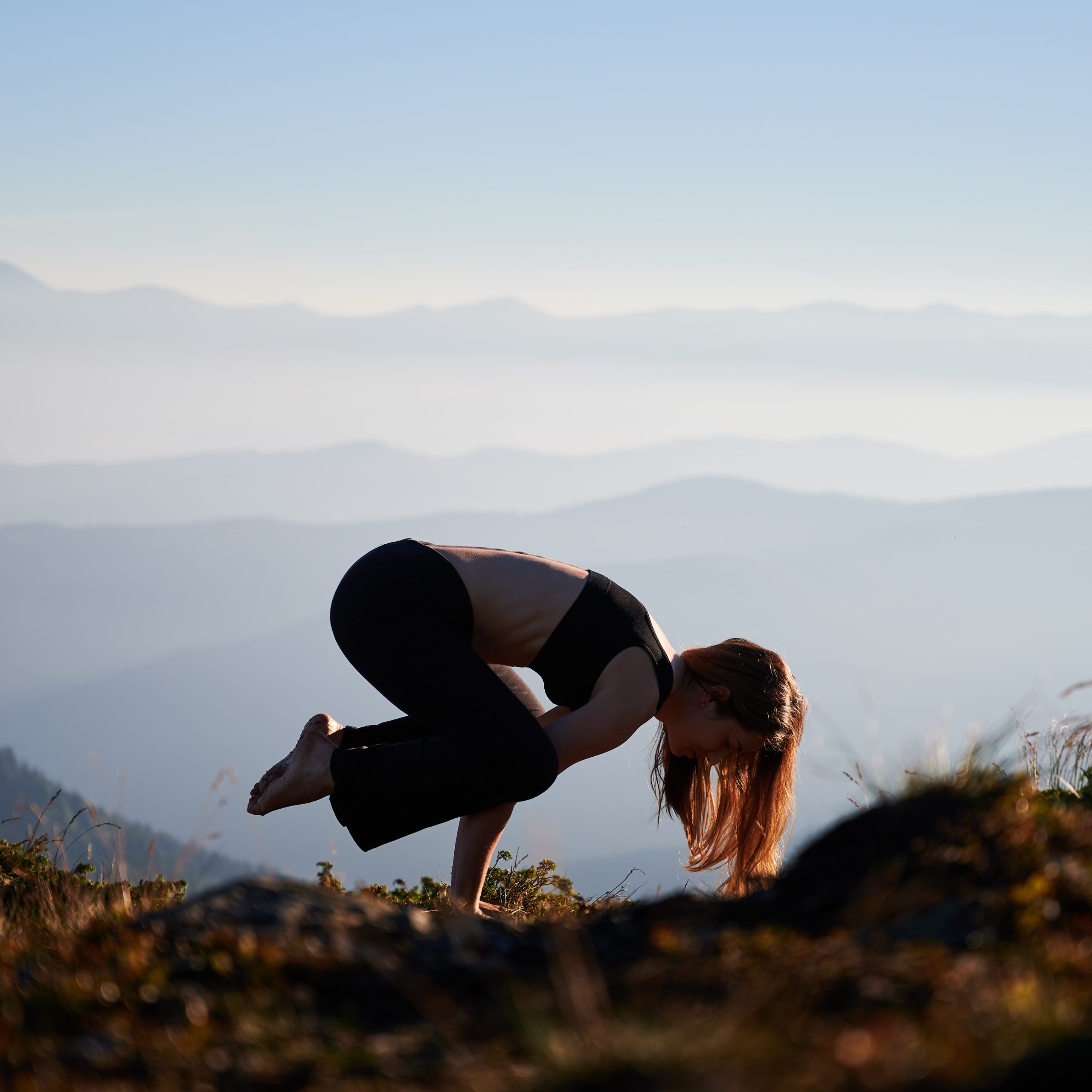 Sporty young woman lifting body and balancing on hands while performing Crow Pose on grassy hill in the morning. Female person practicing yoga outdoors in mountains with blue-white sky on background.